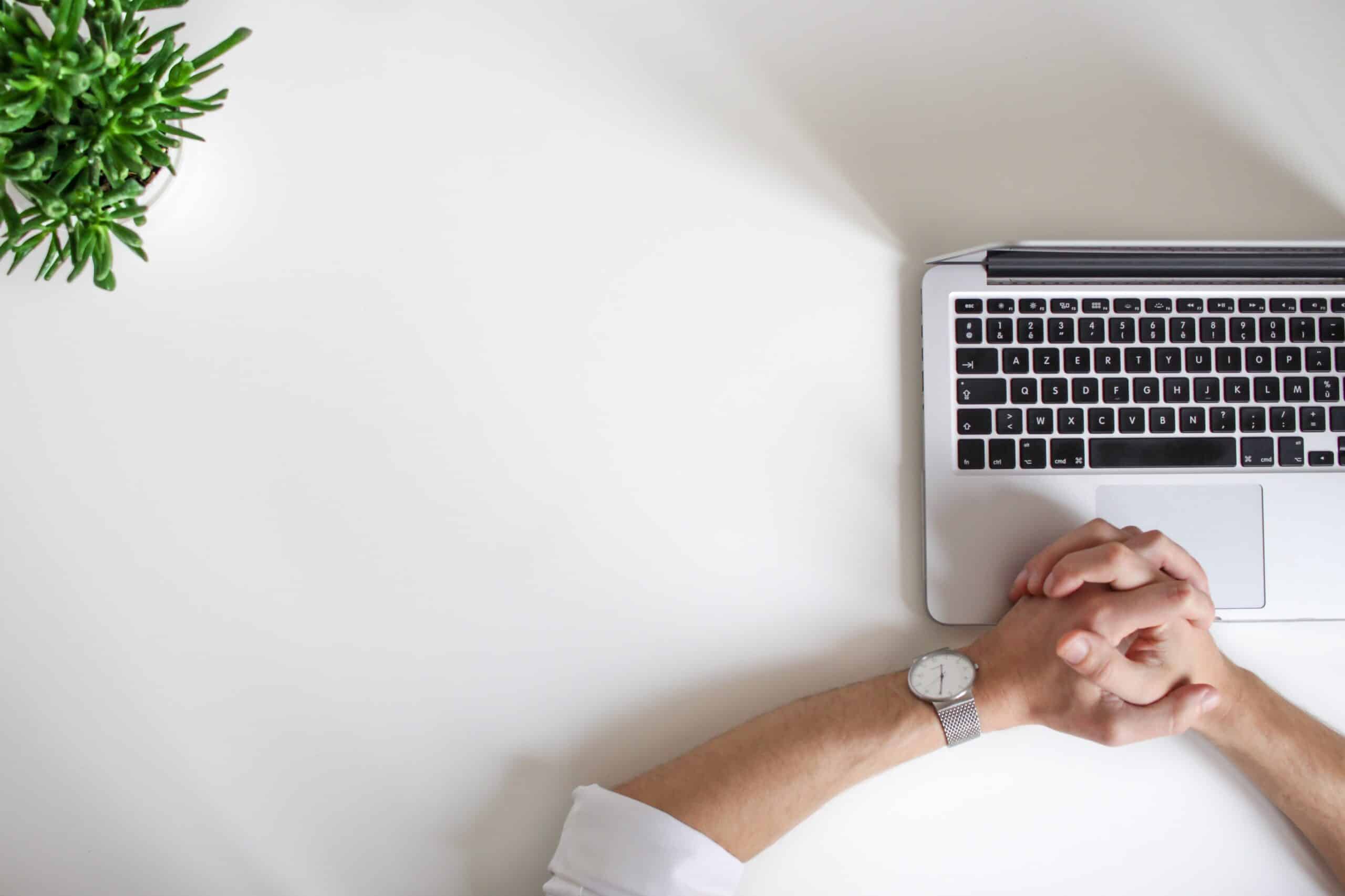 clean white desk with small green plant , crossed hands resting on keyboard